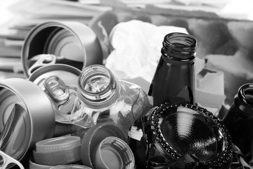 A collection of recyclable household waste items including clear plastic bottles with some caps still attached, several aluminium and plastic drink cans, and a few glass bottles with dark-colored glass, all placed irregularly on a surface. The bottles and cans are surrounded by crumpled tissue paper and other miscellaneous rubbish, possibly in preparation for disposal or recycling. The scene appears to be set outdoors or in a waste collection area, with natural lighting casting soft shadows. The image emphasizes the need for rubbish removal services to handle domestic waste, highlighting common items found in garden clearance or outdoor rubbish disposal tasks. The cluttered arrangement and mixed materials reflect typical household waste that a company like Garden Clearance Kenton may clear from outdoor spaces or gardens in Kenton, helping to keep the environment clean and tidy.
