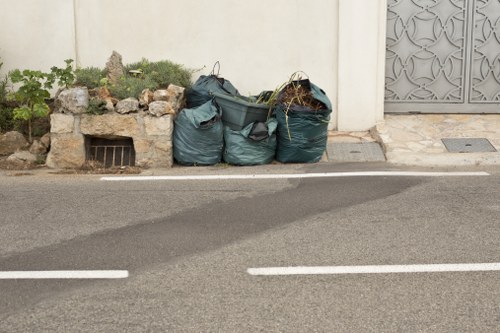 Three large green plastic rubbish bags filled with garden waste are positioned on the edge of a paved road, with a small garden area featuring plants and rocks to the left and a decorative metal gate to the right. The bags appear to contain twigs, leaves, and other garden debris, and are placed near a white wall, possibly the boundary of a residential property. The surface of the road shows a darker strip where vehicles have driven over, and the scene suggests outdoor waste removal services, possibly carried out by Garden Clearance Kenton. The environment is typical of a suburban or residential street, with the garden waste awaiting collection or disposal. The image reflects a clean and organized outdoor setting, with waste ready for removal, illustrating the kind of rubbish management service provided within the Kenton area.