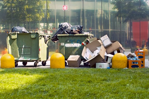 A row of three large wheeled rubbish bins positioned on a paved area adjacent to a building with reflective glass windows, which mirror trees and a red telephone booth in the background. The leftmost bin is green and partially covered with a black plastic bag draped over its opening. The middle bin, also green, has a blue sticker but is overflowing with black plastic bags and miscellaneous waste visible at the top. The rightmost bin is facing slightly to the right and is filled with cardboard boxes and discarded packaging, some of which are torn open and stacked haphazardly. Surrounding the bins are several loose cardboard boxes and blue plastic crates, some crumpled or tipped over. In the foreground, a strip of well-maintained green grass separates the waste area from the rest of the pavement, indicating a maintained outdoor space. This scene illustrates outdoor rubbish collection or waste clearance activity, typical of a professional waste removal service such as Garden Clearance Kenton serving Kenton and surrounding areas, focusing on the disposal of bulky refuse and packaging materials.