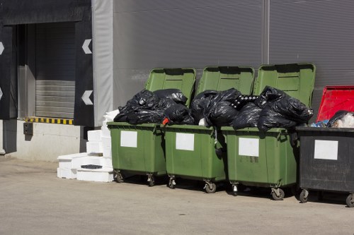 Four green wheeled rubbish bins with open lids are lined up on a paved surface in an industrial or commercial area, with black bin bags filled with waste visible inside the open lids. To the right, a black waste bin with a closed lid is also present. Behind the bins, there is a building facade consisting of a grey roller shutter door and corrugated metal panels, while on the left side, there are concrete blocks stacked behind the bins. The scene appears to be an outdoor waste collection point, typical of a garden or commercial clearance service near Kenton, with clear natural daylight illuminating the area. This setup demonstrates the typical disposal area managed by rubbish removal specialists such as Garden Clearance Kenton, targeting general waste and garden debris collection in the local area.