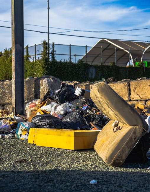 A person wearing a green long-sleeve shirt, a yellow and blue plaid jacket, and light-colored trousers is holding open a large black rubbish bag lined with a shiny black plastic inner layer. The indiv