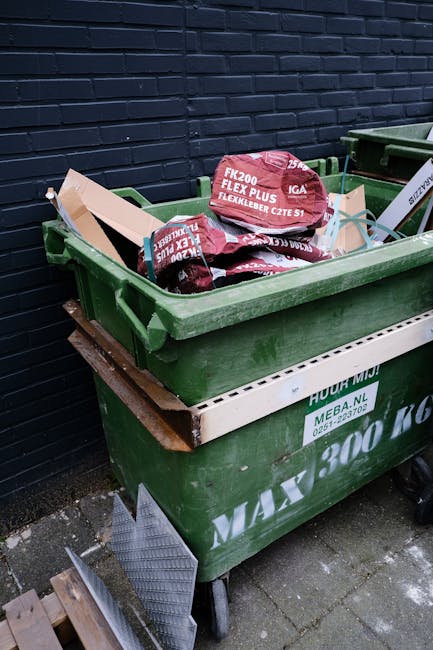 A large green wheeled waste container positioned outdoors on a paved surface, filled with several red and black branded bags containing garden or building waste. The container is placed against a dark