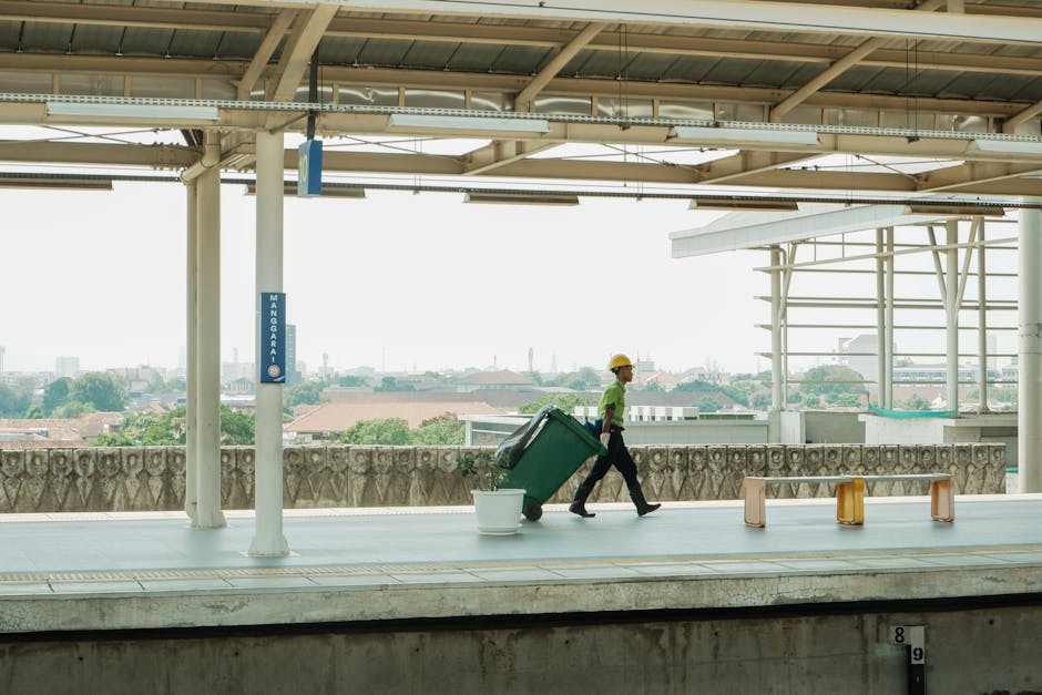 A large collection of mixed household waste and recyclable materials, including paper, cardboard, plastic bags, black and red refuse bins, and assorted packaging, is piled around a metal recycling bin